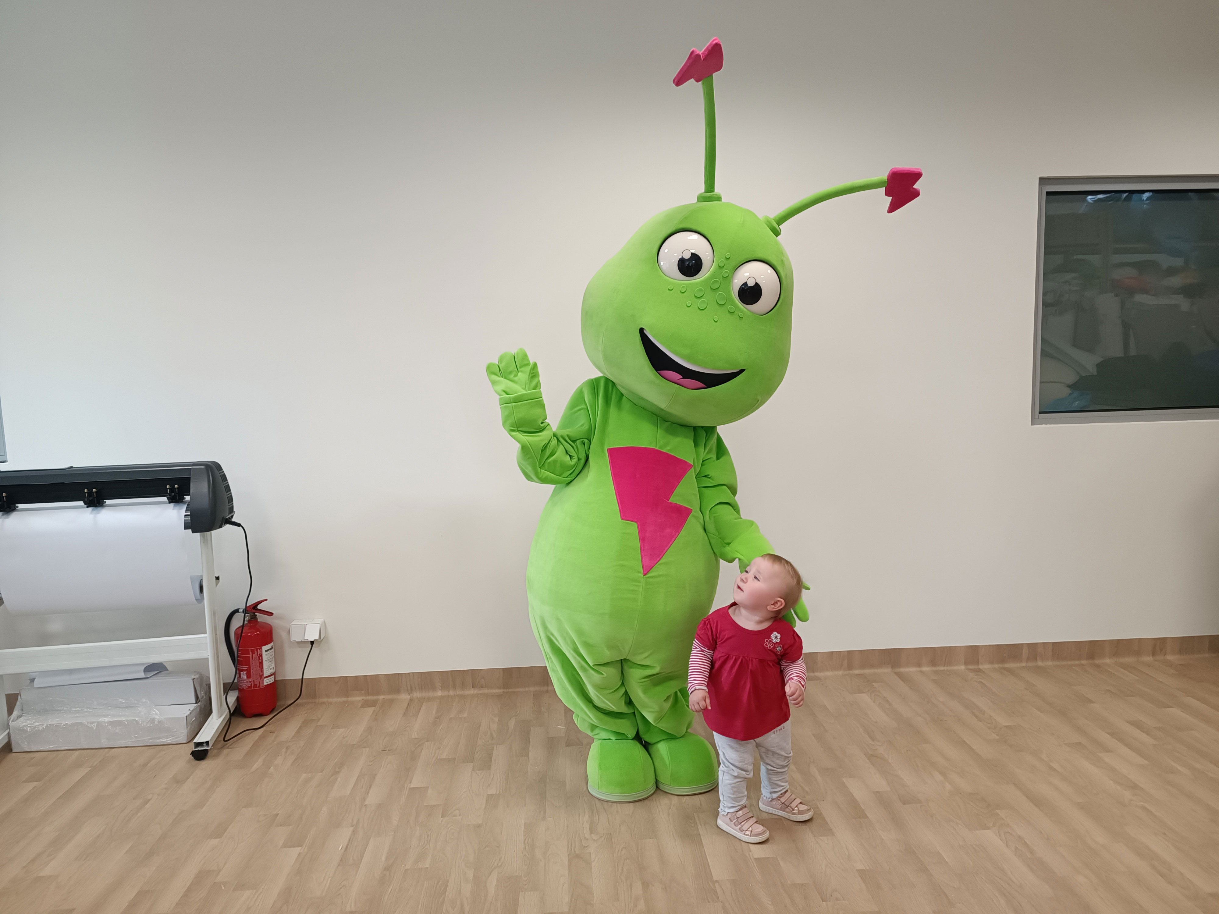 Na fotografii widnieje maskotka w postaci mrówki pozująca do fotografii z liczącą nieco więcej niż 1 rok dziewczynką